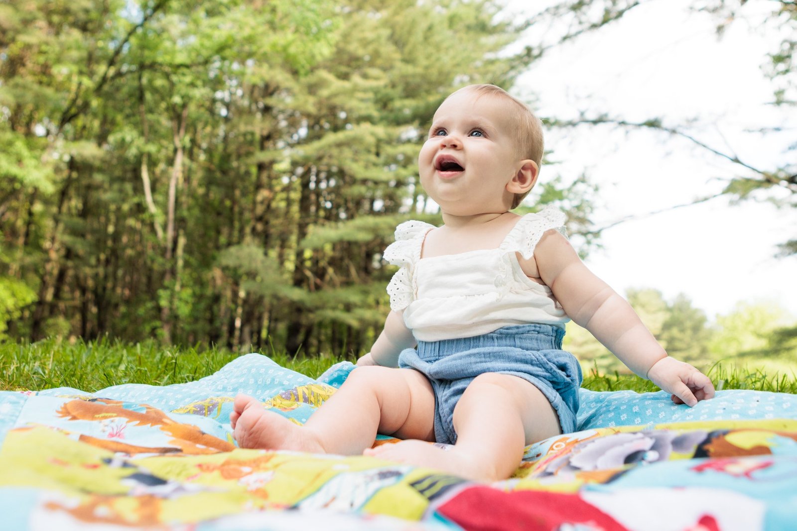 A smiling baby sits on a colorful blanket outdoors